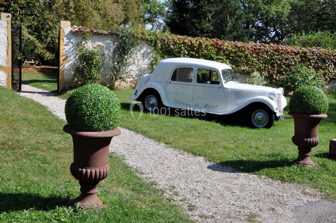 Voiture ancienne blanche stationnée sur une allée gravillonnée, entourée de verdure et de buissons taillés.