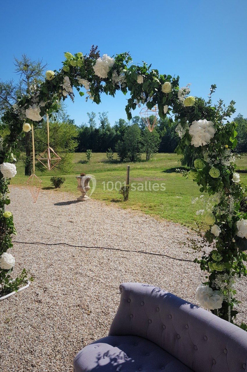 Arche de mariage décorée de fleurs blanches et feuillage, installée sur un chemin gravillonné avec vue sur un jardin.