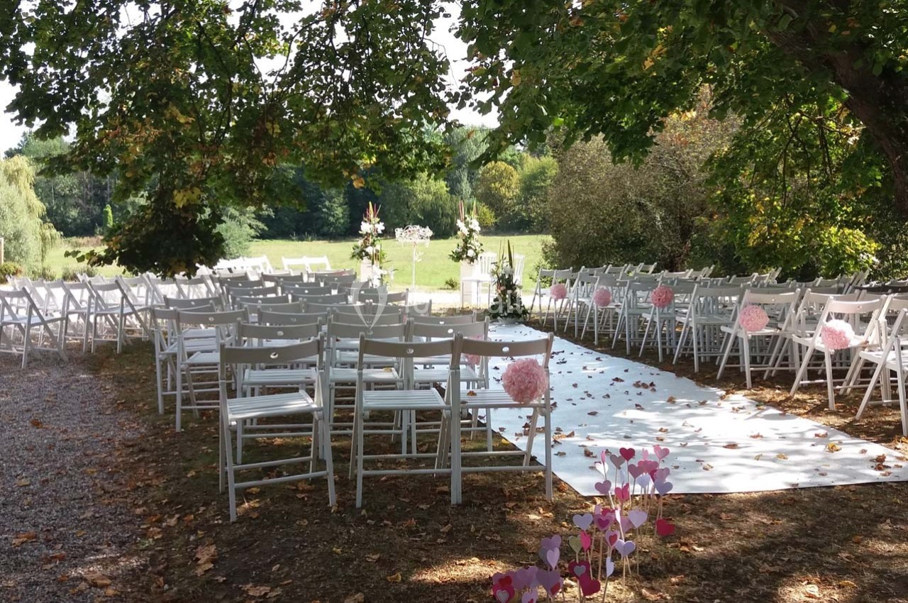 Allée décorée pour une cérémonie en plein air, entourée de chaises blanches et de fleurs roses sous des arbres.