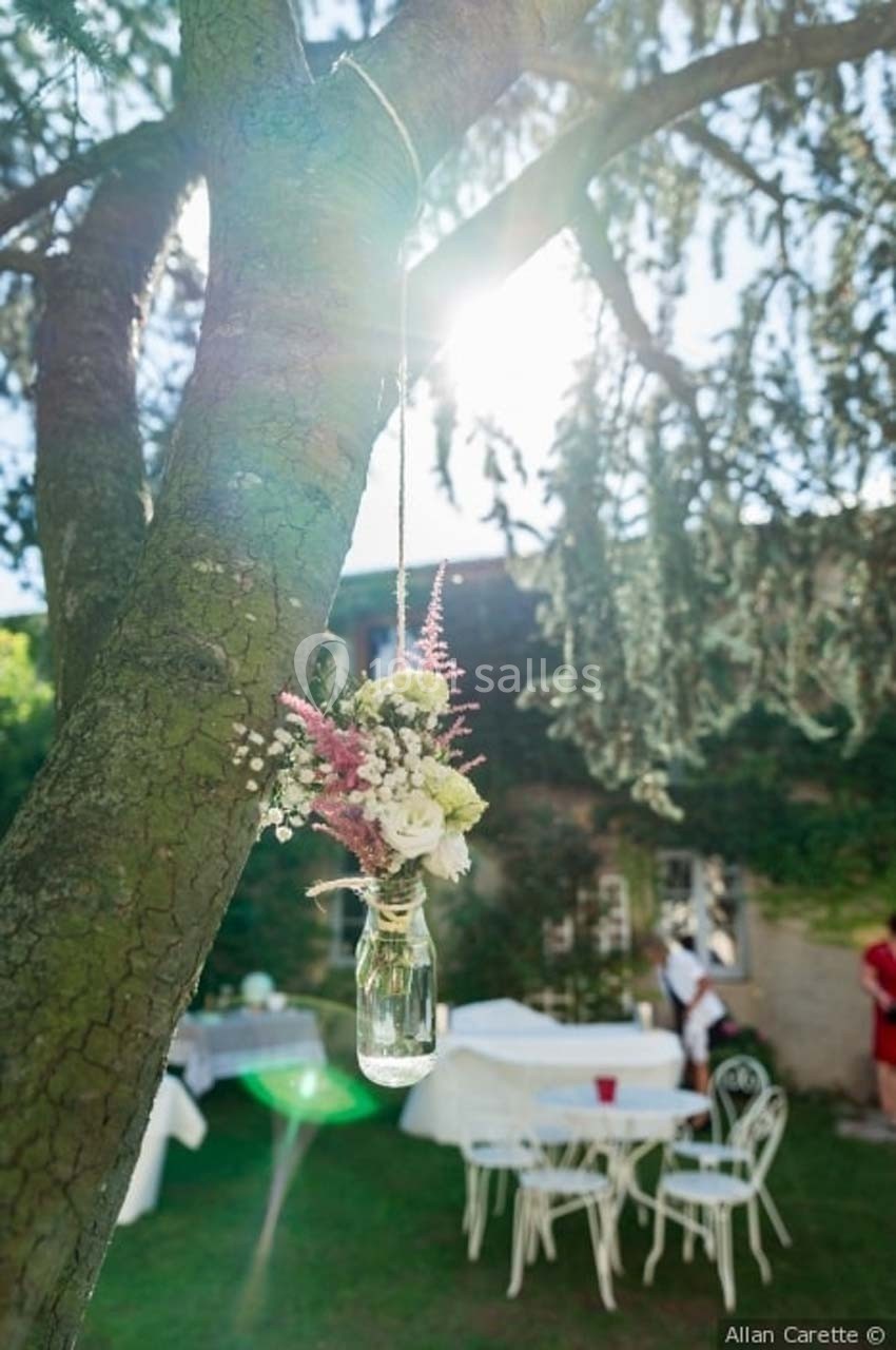 Petit vase en verre suspendu à une branche d'arbre, contenant des fleurs, dans un jardin éclairé par le soleil.