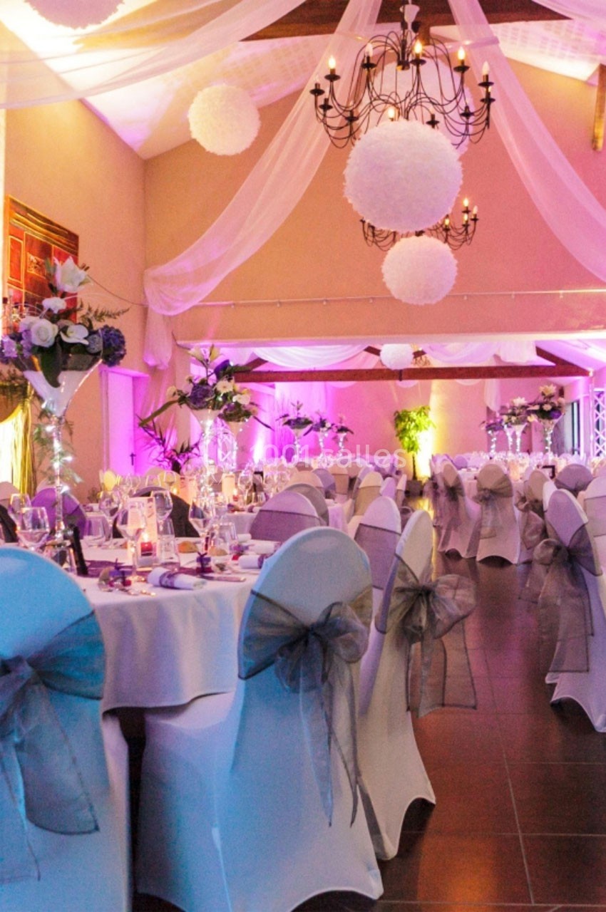 Salle de réception décorée avec des tables dressées, des chaises ornées de nœuds gris et un éclairage rose tamisé.