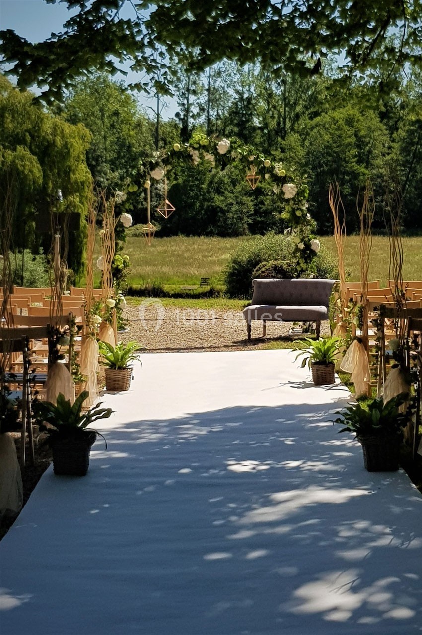 Allée décorée pour une cérémonie en plein air, bordée de plantes et menant à une arche florale devant un champ.