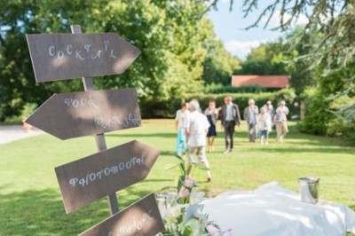 Allée gravillonnée bordée de lanternes blanches menant à un jardin arboré, vue depuis un portail en fer forgé.