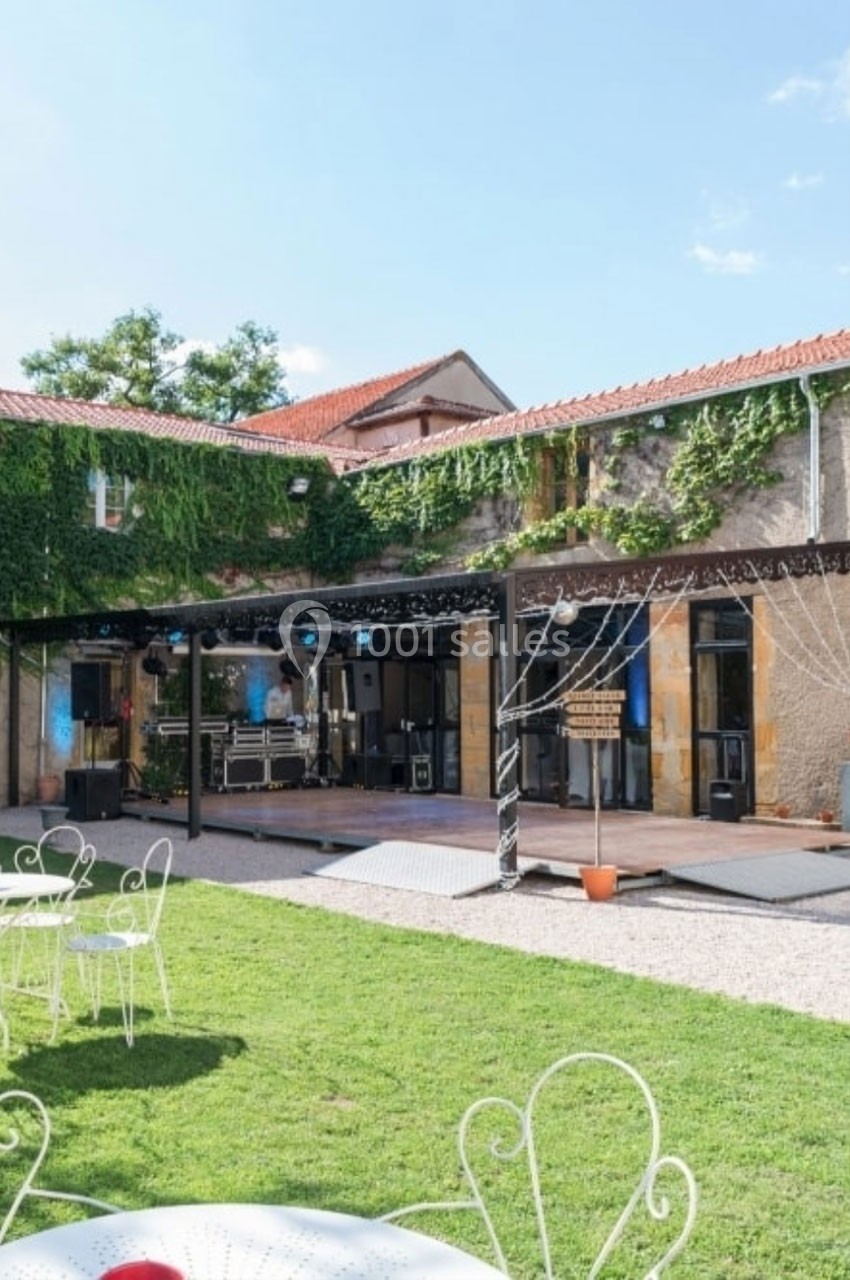 Cour extérieure avec pelouse, mobilier de jardin blanc, scène équipée pour un événement, bâtiment en pierre et lierre.