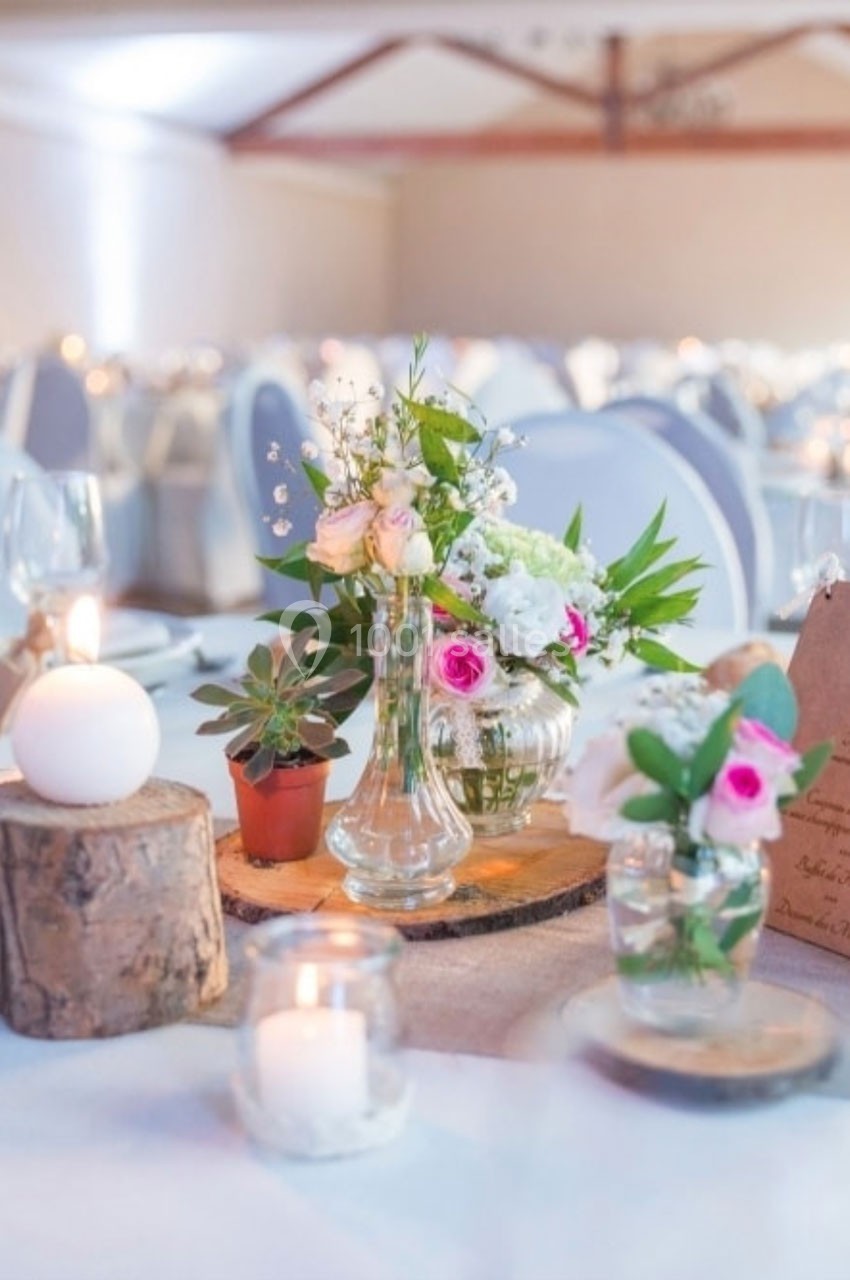 Décoration de table avec bouquets de fleurs, bougies et rondins de bois dans une salle de réception lumineuse.