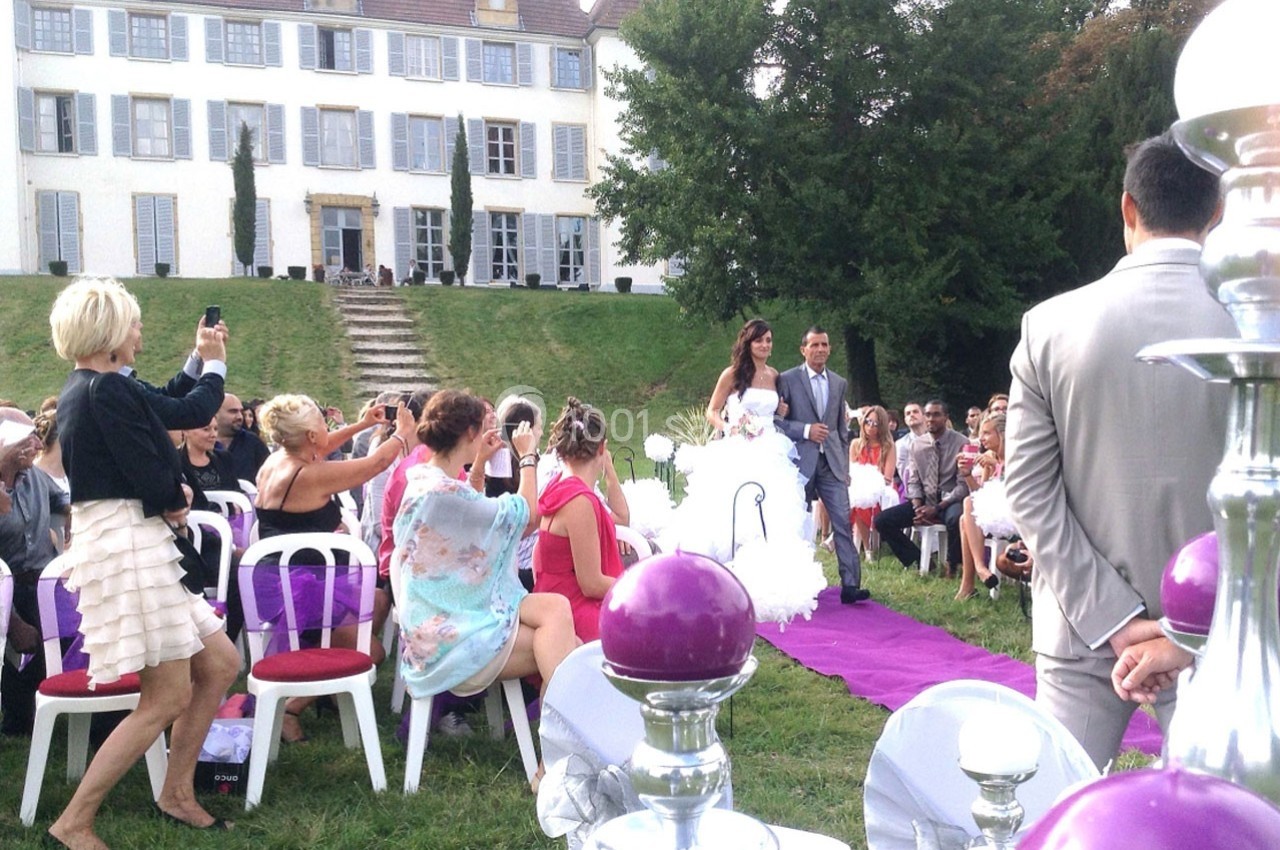 Un couple marche sur un tapis violet lors d'une cérémonie de mariage en plein air devant une grande maison.