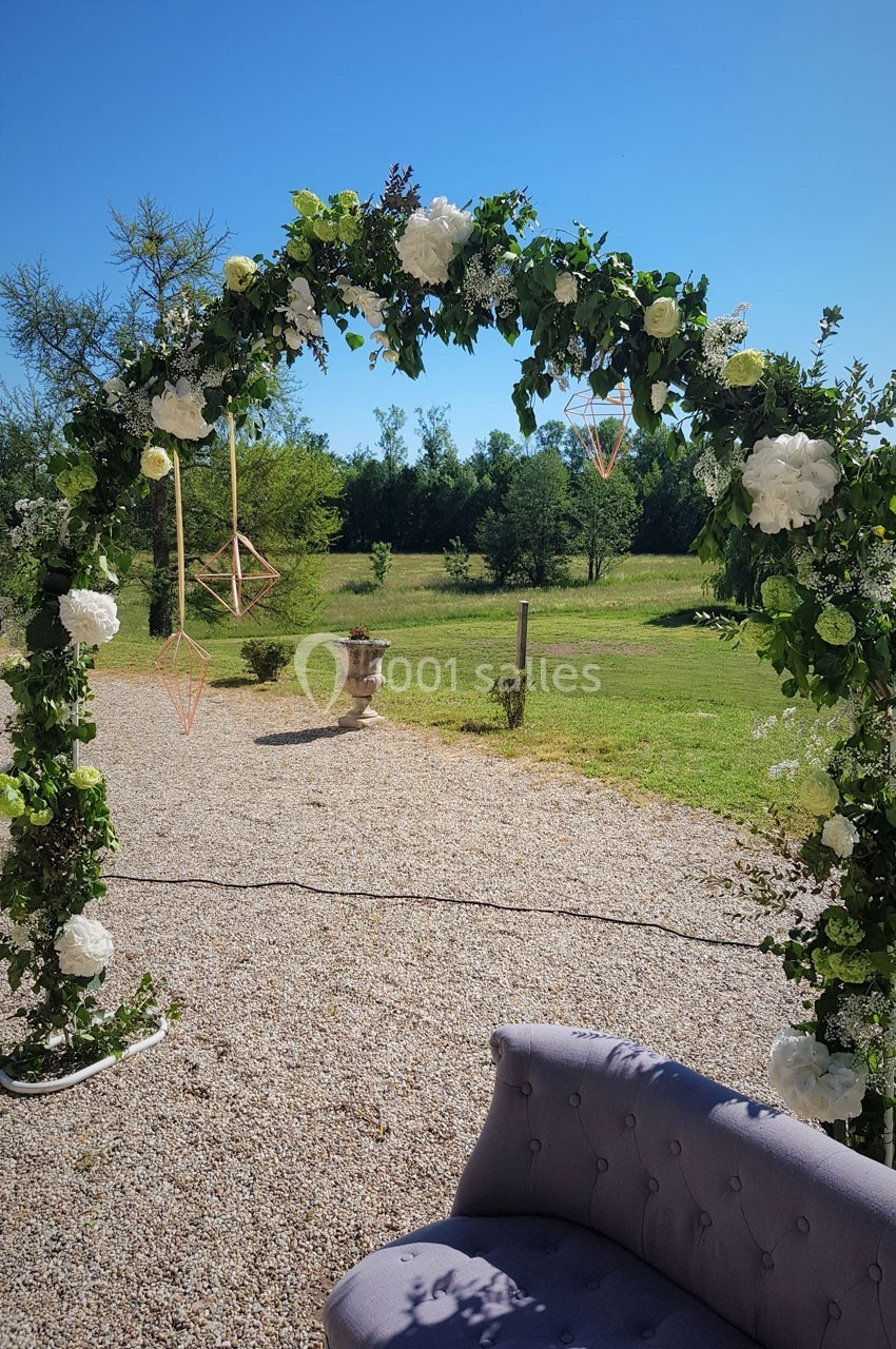 Arche fleurie décorée de feuillage et de fleurs blanches, installée en extérieur sur un sol gravillonné.