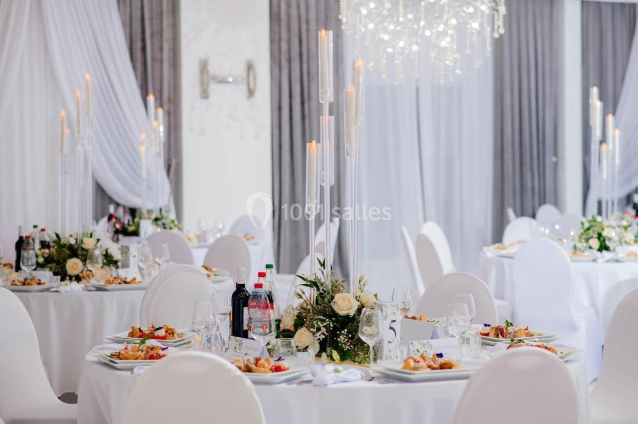 Salle de réception élégante avec tables rondes dressées, décorations florales et chandeliers, ambiance lumineuse.