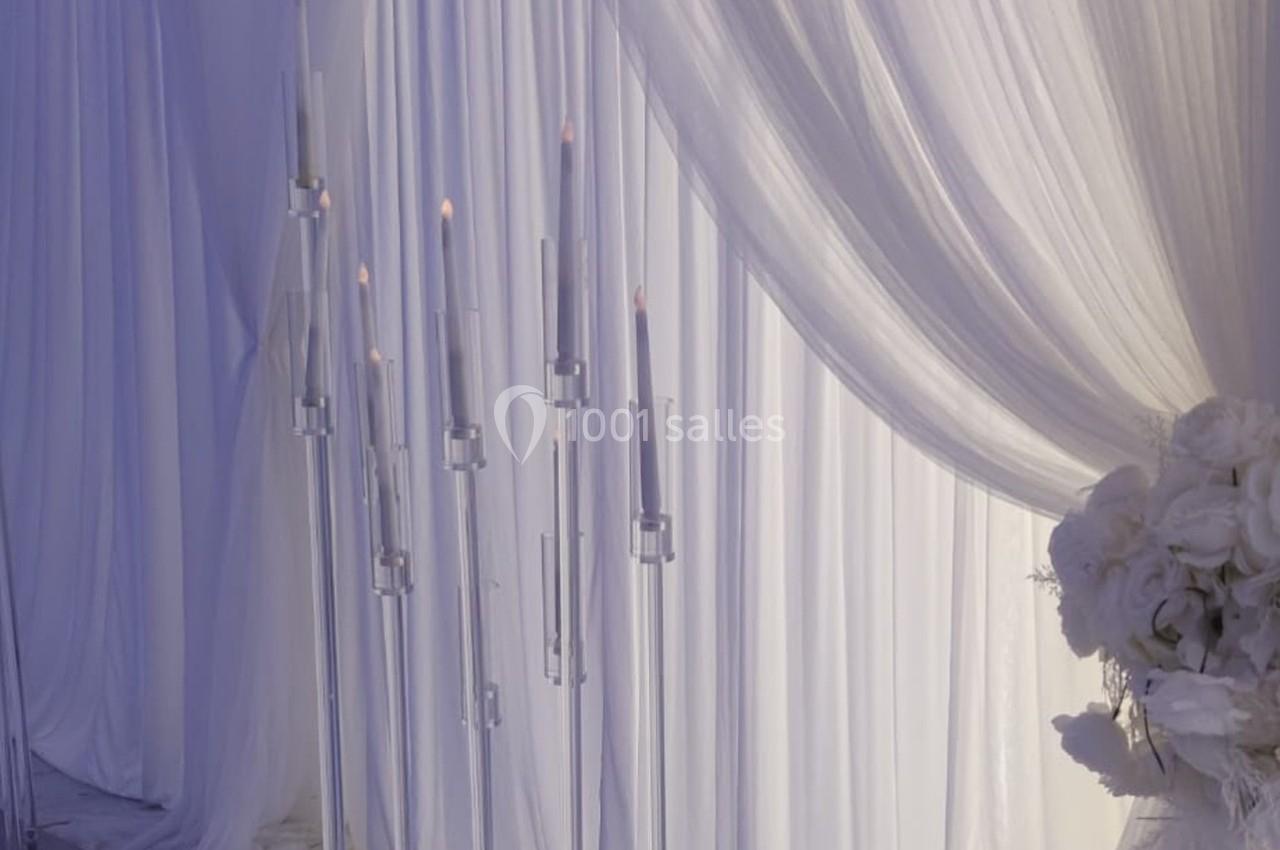 Bougies allumées sur des supports en verre devant des rideaux blancs drapés, avec des fleurs blanches à droite.