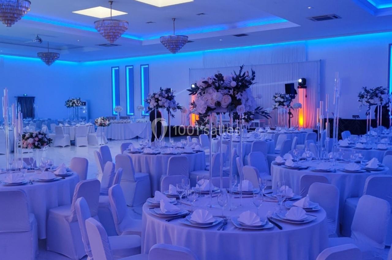 Salle de réception élégante avec tables rondes dressées, décorations florales et éclairage bleu tamisé.