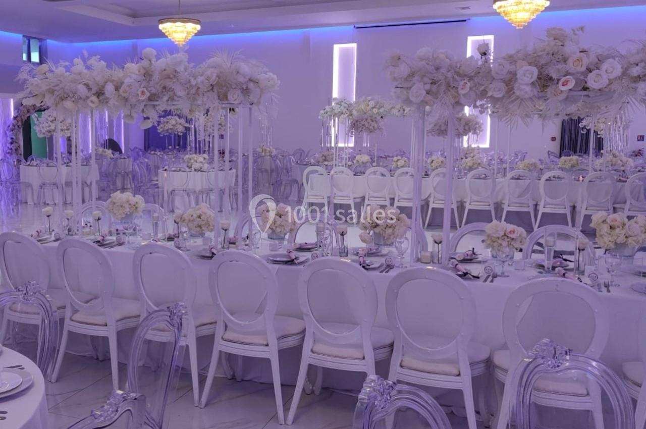 Salle de réception décorée avec des tables blanches, chaises transparentes et arrangements floraux blancs élégants.