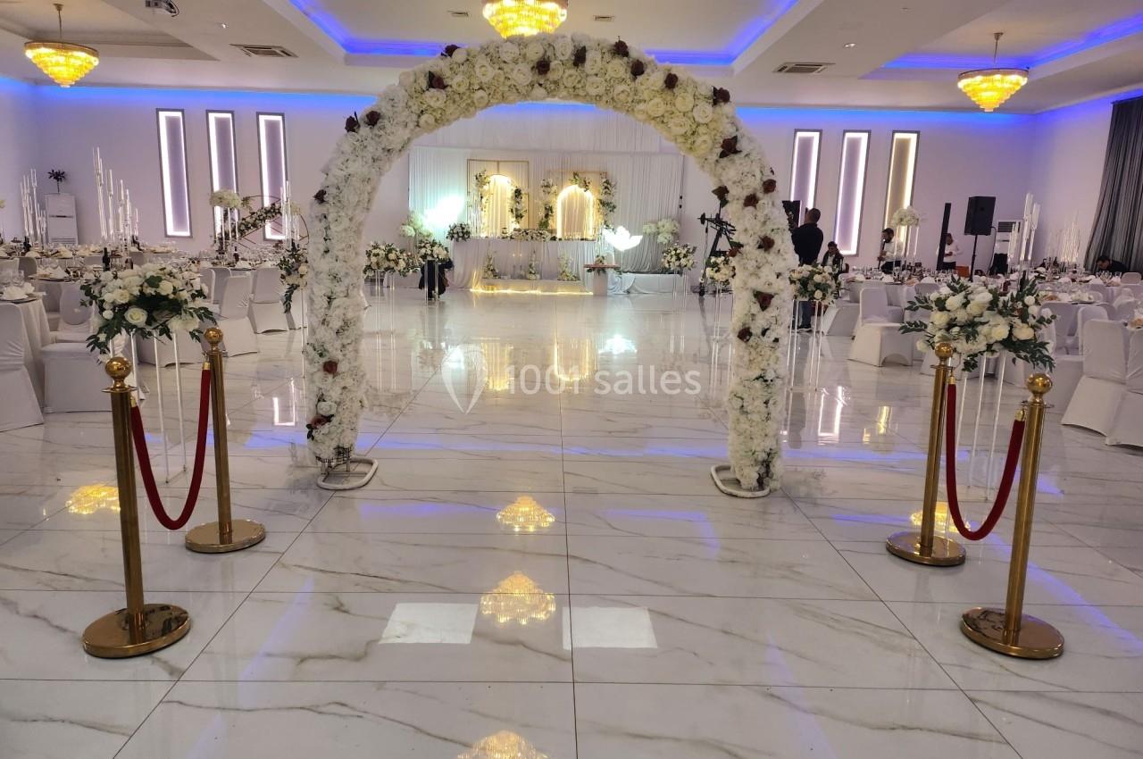 Salle de réception élégante avec un arc floral blanc, des tables décorées et un éclairage lumineux.