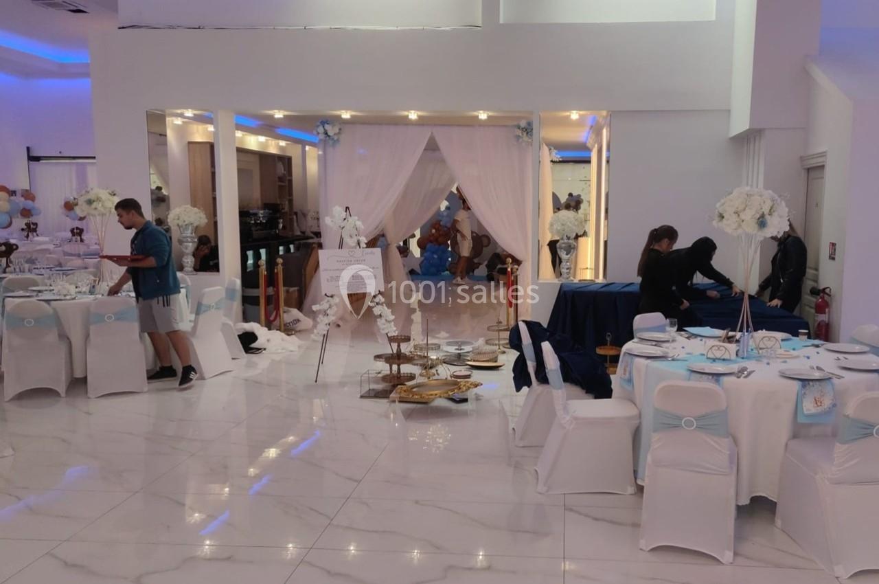 Salle de réception décorée avec des tables dressées, des fleurs blanches et des rideaux blancs encadrant une entrée.