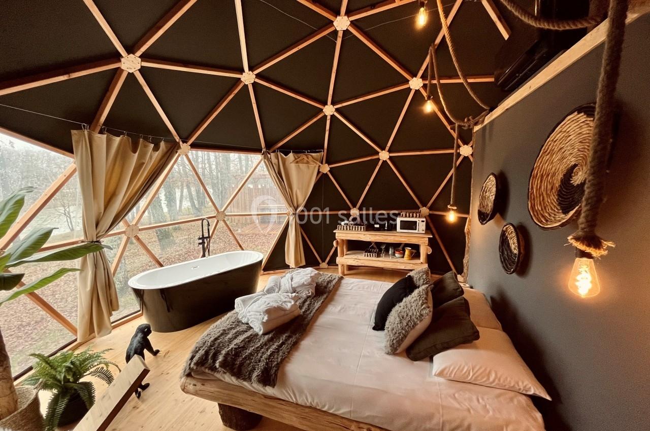 Intérieur d'un dôme géodésique avec lit en bois, baignoire, rideaux ouverts sur un paysage boisé et décoration naturelle.