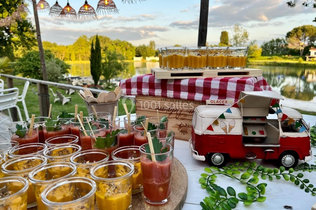 Buffet en extérieur avec verrines colorées, nappe à carreaux rouges et camionnette miniature près d'un étang.