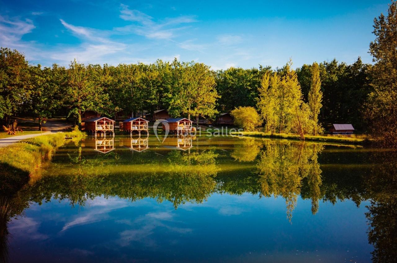 Chalets en bois alignés au bord d'un lac calme, entourés d'arbres et reflétés dans l'eau sous un ciel bleu.