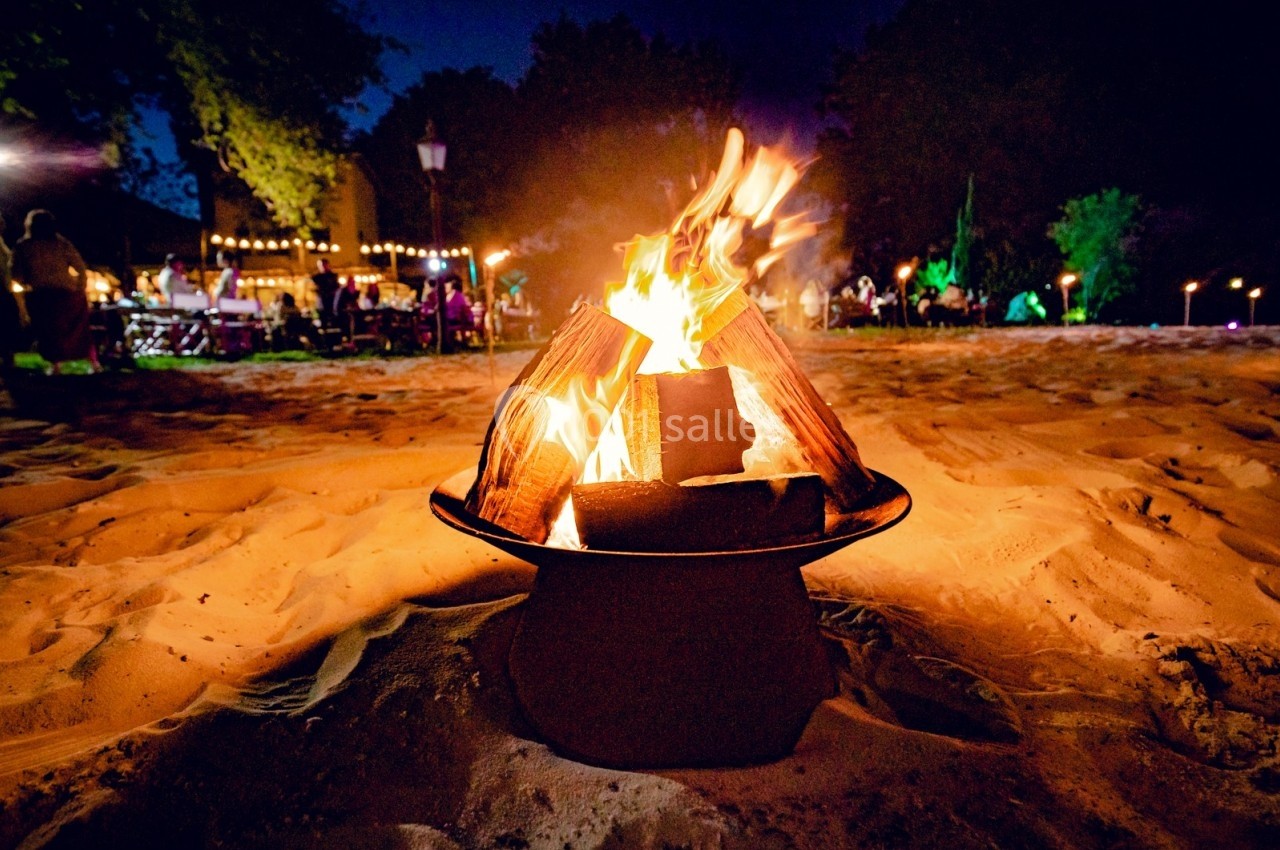 Feu de camp allumé sur du sable, entouré de lumières et de personnes en arrière-plan dans une ambiance nocturne.