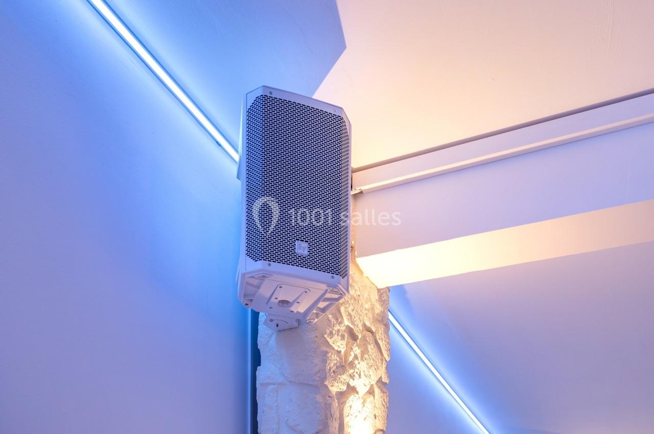 Enceinte murale blanche fixée sur un mur en pierre, éclairée par une lumière bleue et blanche.