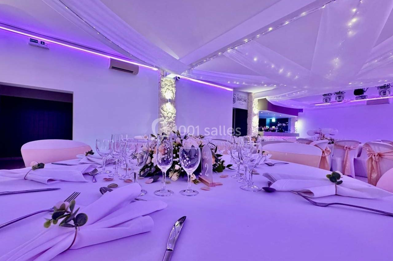 Salle de réception élégamment décorée avec tables dressées, nappes blanches, verres et éclairage violet tamisé.