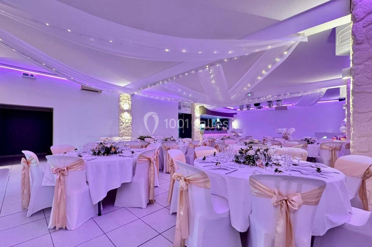 Salle de réception décorée avec des tables rondes, nappes blanches, chaises ornées de rubans et éclairage violet.