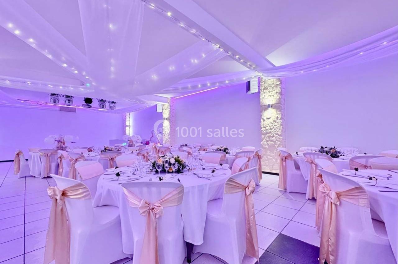 Salle de réception décorée avec des tables rondes, nappes blanches, chaises ornées de nœuds roses et éclairage tamisé.