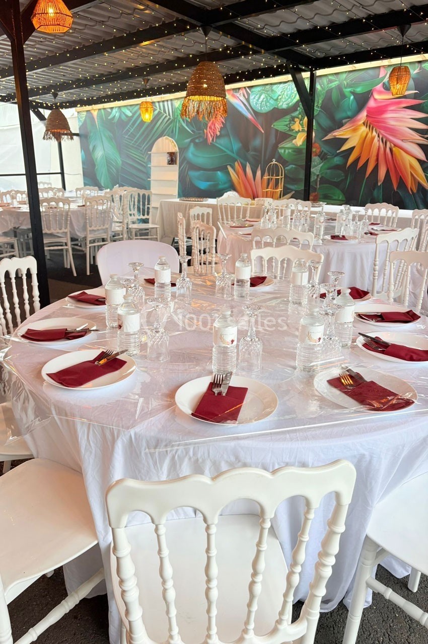 Tables rondes dressées avec nappes blanches, serviettes rouges et vaisselle, dans une salle décorée de motifs tropicaux.