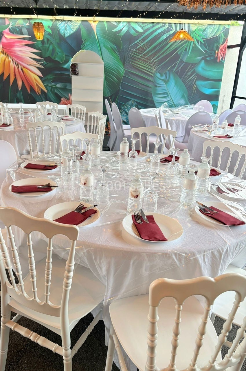 Tables rondes dressées avec nappes blanches, serviettes bordeaux et chaises blanches, décor mural tropical en arrière-plan.