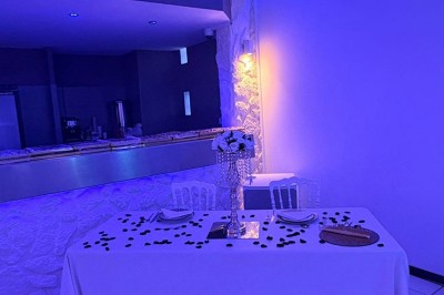 Salle de réception décorée avec des tables rondes, nappes blanches, chaises élégantes et éclairage violet tamisé.