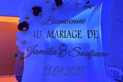 Salle de réception décorée avec des tables rondes, nappes blanches, chaises élégantes et éclairage violet tamisé.
