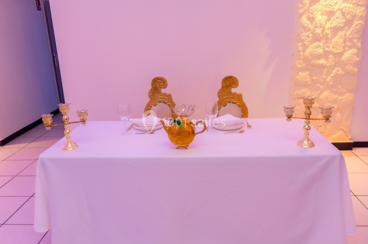Table décorée avec nappes blanches, chaises dorées, chandeliers et une théière centrale dans un cadre lumineux.