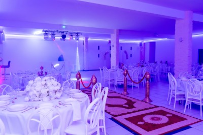 Salle de réception décorée avec des tables rondes, nappes blanches, chaises élégantes et éclairage violet tamisé.