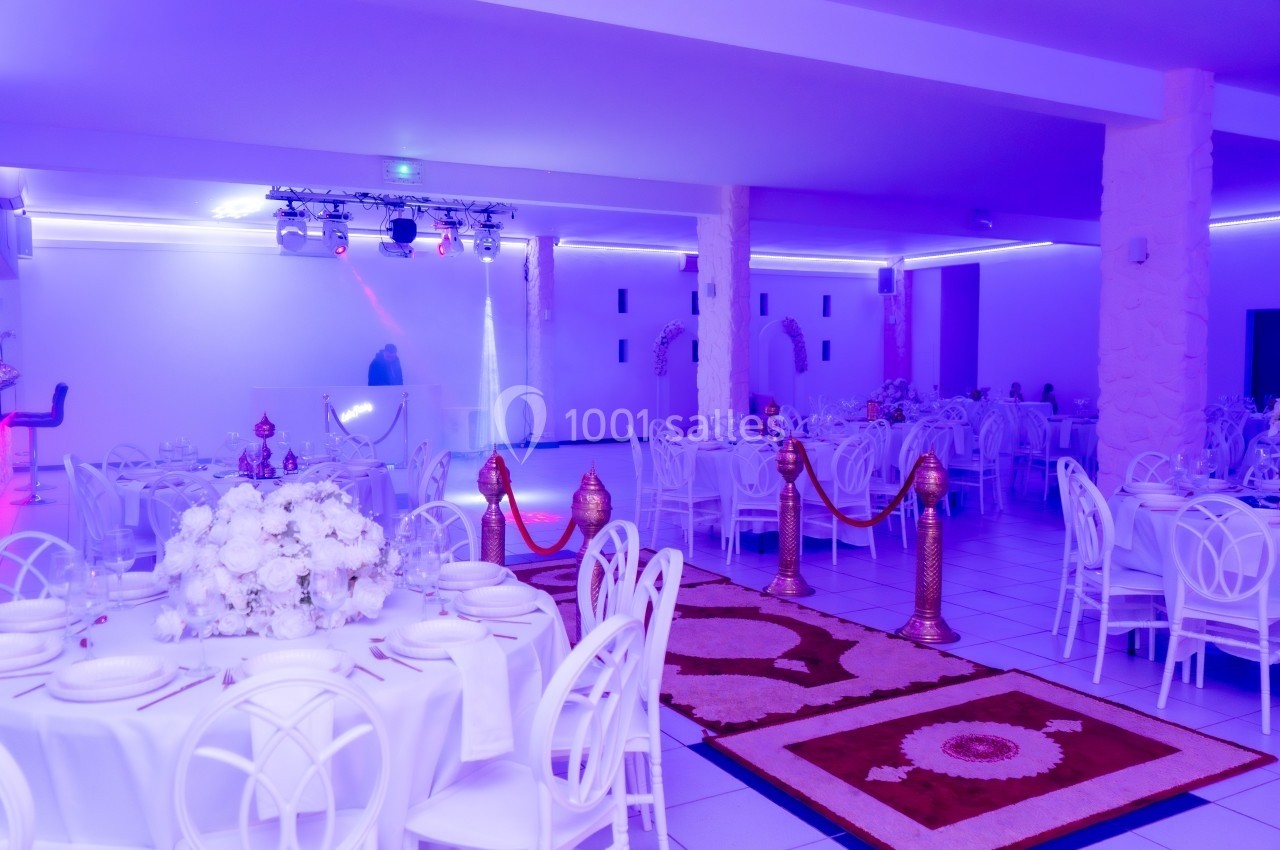 Salle de réception décorée avec des tables dressées, des fleurs blanches et un éclairage violet.