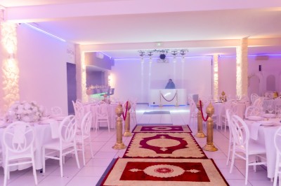 Salle de réception décorée avec des tables rondes, nappes blanches, chaises élégantes et éclairage violet tamisé.