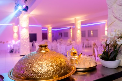 Salle de réception décorée avec des tables rondes, nappes blanches, chaises élégantes et éclairage violet tamisé.