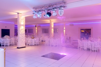 Salle de réception décorée avec des tables rondes, nappes blanches, chaises élégantes et éclairage violet tamisé.
