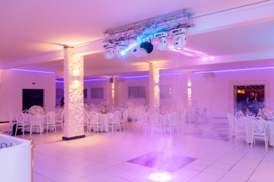 Salle de réception décorée avec des tables rondes, nappes blanches, chaises élégantes et éclairage violet tamisé.