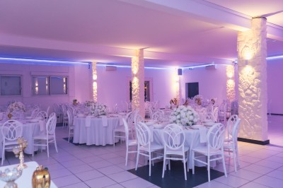 Salle de réception décorée avec des tables rondes, nappes blanches, chaises élégantes et éclairage violet tamisé.