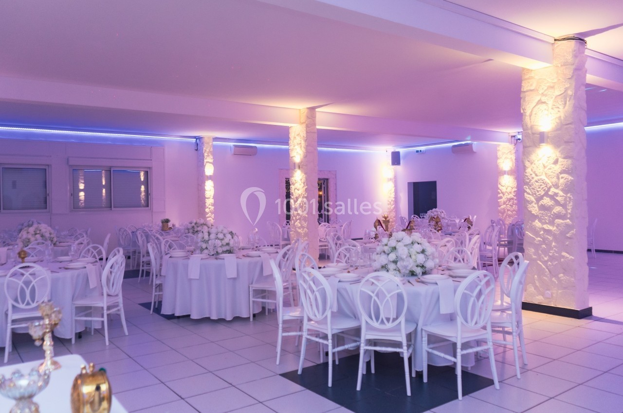 Salle de réception lumineuse avec tables rondes décorées de nappes blanches et bouquets de fleurs.