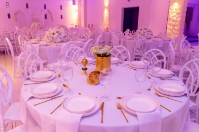 Salle de réception décorée avec des tables rondes, nappes blanches, chaises élégantes et éclairage violet tamisé.