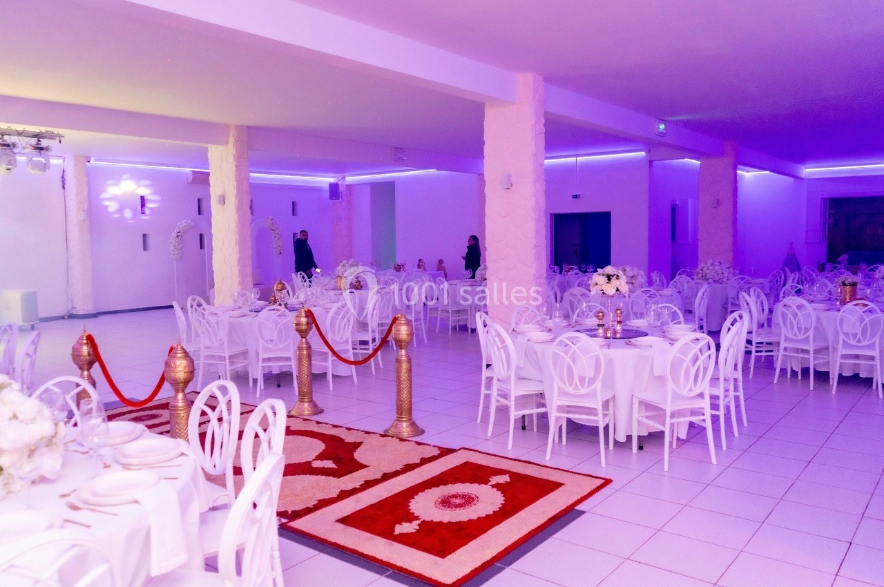 Salle de réception lumineuse avec tables rondes dressées, chaises blanches et tapis rouges délimitant un espace central.