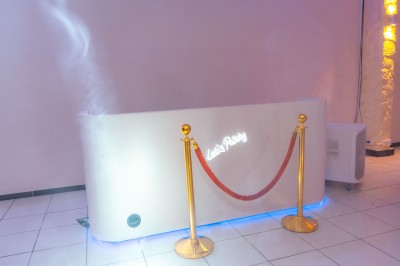 Salle de réception décorée avec des tables rondes, nappes blanches, chaises élégantes et éclairage violet tamisé.