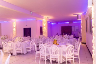 Salle de réception décorée avec des tables rondes, nappes blanches, chaises élégantes et éclairage violet tamisé.