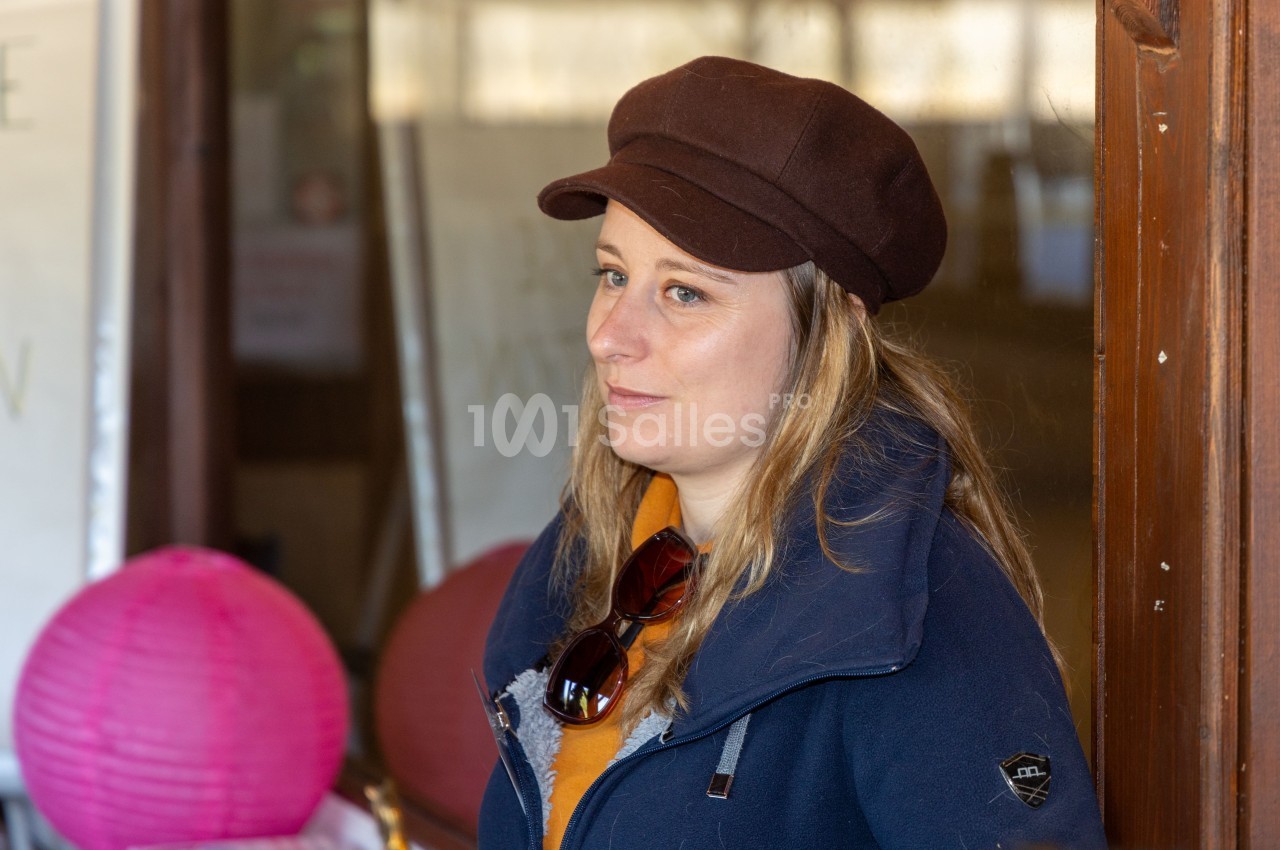 Femme portant une casquette marron et une veste bleue, debout près d'une porte en bois avec des lanternes roses en arrière…