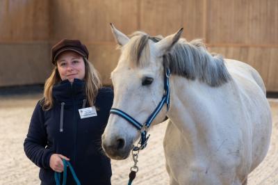 Femme souriante tenant les rênes d'un cheval brun dans un environnement extérieur boisé.