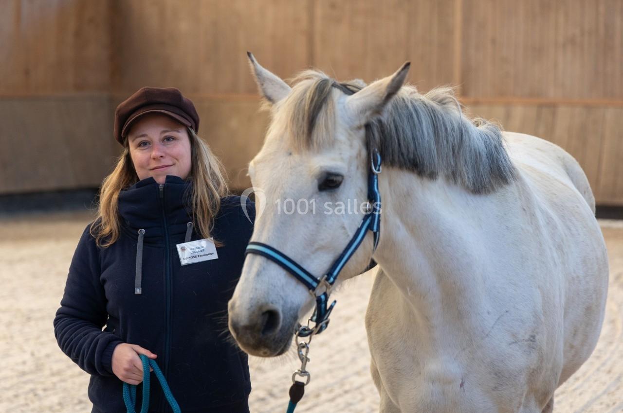 Une femme souriante tient un cheval blanc en licol bleu dans un manège équestre en bois.