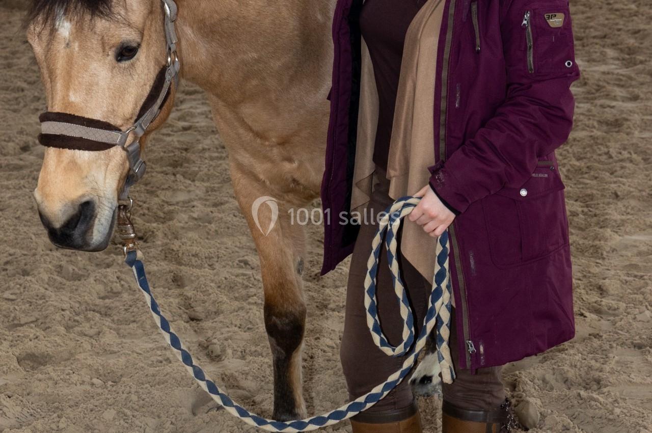 Une femme souriante tient un cheval par une longe dans un manège intérieur sur un sol sablonneux.