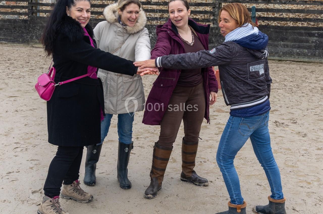 Quatre femmes souriantes se tiennent la main en cercle dans une cour en terre, vêtues de manteaux d'hiver.