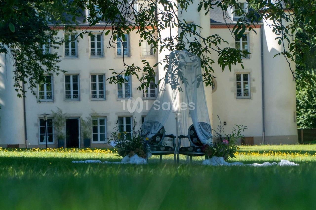Deux chaises longues avec un voile blanc, entourées de fleurs, installées sur une pelouse devant un bâtiment ancien.