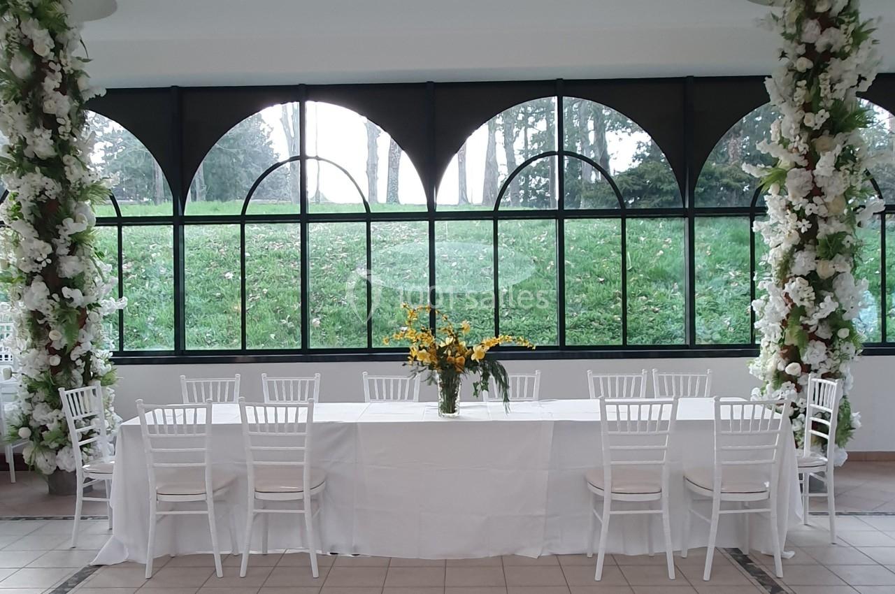 Salle lumineuse avec une grande table blanche décorée de fleurs jaunes, entourée de chaises blanches et de colonnes fleuries.