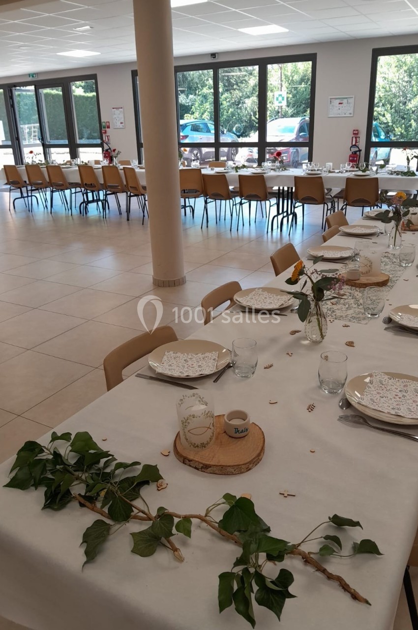 Salle de réception avec tables dressées, décorées de feuillages et vaisselle, éclairée par de grandes baies vitrées.